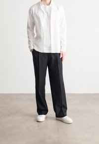 Man in een losse zwarte broek, witte sneakers, wit T-shirt en een open, witte lange mouwen shirt, staande op een lichtgrijze vloer.