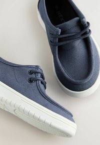 Marineblaue Segelschuhe aus Canvas mit weißen Gummisohlen, ausgestattet mit zwei Ösen und einem glatten, abgerundeten Zehenbereich. Strukturiertes Obermaterial.