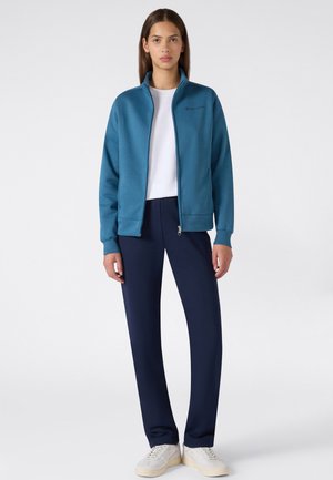 Mujer de pie con chaqueta turquesa con cremallera, camisa blanca, pantalones azul marino y zapatillas blancas sobre un fondo claro y liso.