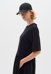 Černé šaty s krátkým rukávem a uvolněným střihem, se splývavou sukní a kapsami. Nosit s černou baseballovou čepicí, minimalistický design.