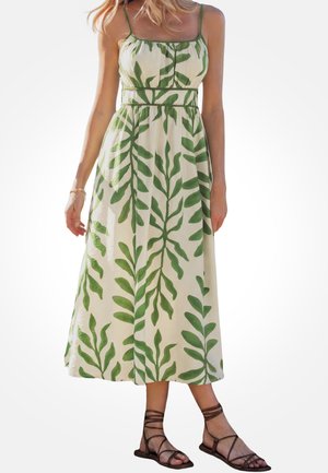Vrouw draagt een mouwloze crèmekleurige jurk met groene bladpatronen, gecombineerd met bruine sandalen met bandjes en platte zolen, staand tegen een effen achtergrond.