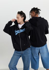 Sort zip-up hoodie med et blåt grafisk design på forsiden, båret af to personer, der vender i modsat retning. Lyseblå jeans og shorts.