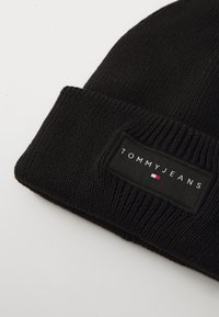 Black knit beanie featuring a rectangular fabric label with "TOMMY JEANS" text and a small red, white, and blue flag emblem.