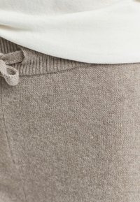 Nærbilde av beige strikkede bukser med snøring i midjen og en hvit topp som er stukket inn, som viser teksturert stoff og sømdetaljer.