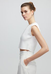 Witte cropped top met korte mouwen, voorzien van een rits aan de voorkant en decoratieve afwerking. Gecombineerd met hoge witte broeken, wat zorgt voor een strakke uitstraling.