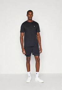 Schwarzes T-Shirt mit kurzen Ärmeln und schwarze Shorts, beide aus glattem Stoff. Weißes Logo auf der linken Brustseite und an der Seite der Shorts. Weiße Turnschuhe.