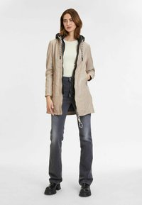Beige hooded jacket met een gladde afwerking, met een rits voorkant en zwarte accenten. Gecombineerd met grijze jeans en zwarte schoenen.