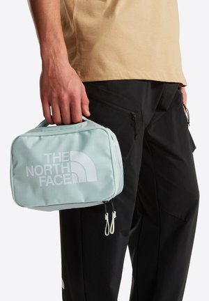 Άτομο που φοράει μαύρο παντελόνι και μπεζ πουκάμισο κρατώντας μια ανοιχτό μπλε ορθογώνια θήκη North Face από τη λαβή της.