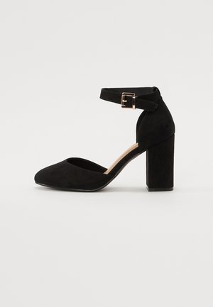 Czarny czółenek na kostkę z blokowym obcasem, zamszową fakturą, spiczastym noskiem i małym metalowym zapięciem na pasku.