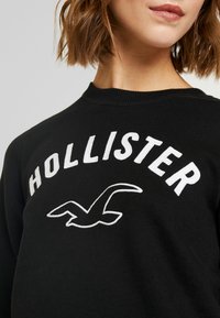Černá mikina s bílým vyšitým textem "HOLLISTER" a logem racka. Materiál vypadá měkký s klasickým kulatým výstřihem.