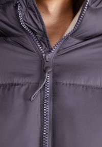 Dunkelgraue Steppjacke mit hohem Kragen, sichtbarem Reißverschluss mit strukturiertem Reißverschlussgriff und glatter, glänzender Stoffoberfläche.