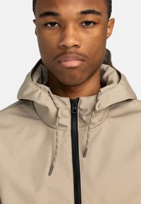 Beige Kapuzenjacke mit hohem Kragen, Frontreißverschluss und verstellbaren Zugbändern. Glatte Textur und kontrastierender schwarzer Reißverschluss.