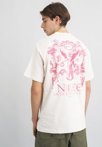 Κρέμα βαμβακερή μπλούζα με μεγάλο ροζ γραφικό σχέδιο στην πλάτη ενός αγγέλου και κείμενο, με χαλαρή εφαρμογή και κοντά μανίκια.