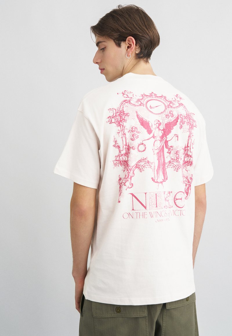 Κρέμα βαμβακερή μπλούζα με μεγάλο ροζ γραφικό σχέδιο στην πλάτη ενός αγγέλου και κείμενο, με χαλαρή εφαρμογή και κοντά μανίκια.