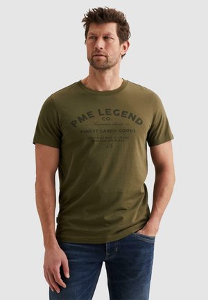 Man draagt een olijfgroen PME Legend t-shirt met zwarte bedrukte tekst, staat met één hand in de zak en kijkt opzij.