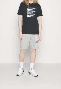 Person som bär en svart Nike T-shirt med tre swoosh-logotyper, grå Nike-shorts med flera logotyper, vita strumpor och vita Nike-sneakers.