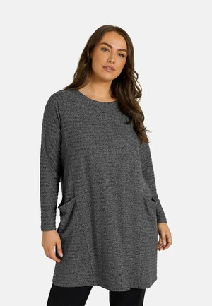 Vrouw met lang bruin haar die een getextureerde donkergrijze tuniek met lange mouwen en zakken aan de voorkant draagt, staand tegen een effen lichte achtergrond.