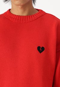 Pull rouge en tricot avec col côtelé, arborant un motif de cœur noir brisé sur le côté gauche de la poitrine. Texture lisse, coupe décontractée.