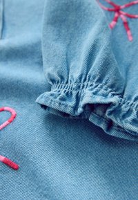 Lys blå denimstof med gatherede ærmer. Har pink broderede accenter i kurvede designs. Blød tekstur med synlig vævemønster.