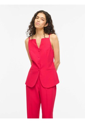 Kobieta o kręconych ciemnych włosach ubrana w jasnoczerwony bezrękawnik z guziczkami i pasujące szerokie spodnie na białym tle.