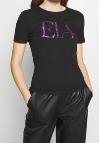 Svart kortärmad t-shirt i bomullsblandning med lila paljetterad "EA" logotyp. Fitted design med rund halsringning, kombinerad med svarta läderbyxor.