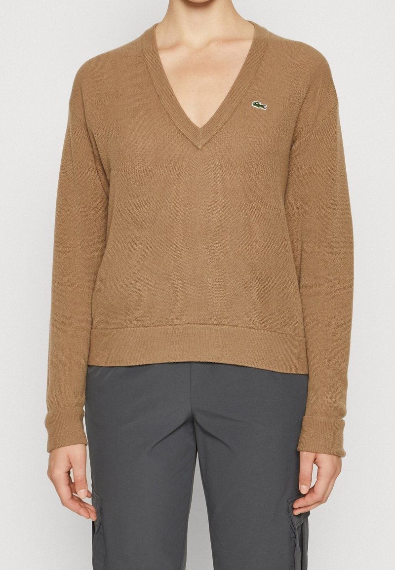 Brun v-ringad tröja i mjukt, texturerat stickat material, med ribbad nederkant och ärmslut, samt en liten grön logotyp på vänster bröst.