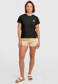 Black cotton t-shirt with short sleeves, round neckline, and a small circular logo on the chest. Paired with beige textured shorts and white sandals.