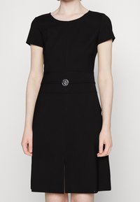 Woman wearing a knee-length black dress with short sleeves and a round neckline, featuring a decorative silver circular belt buckle.