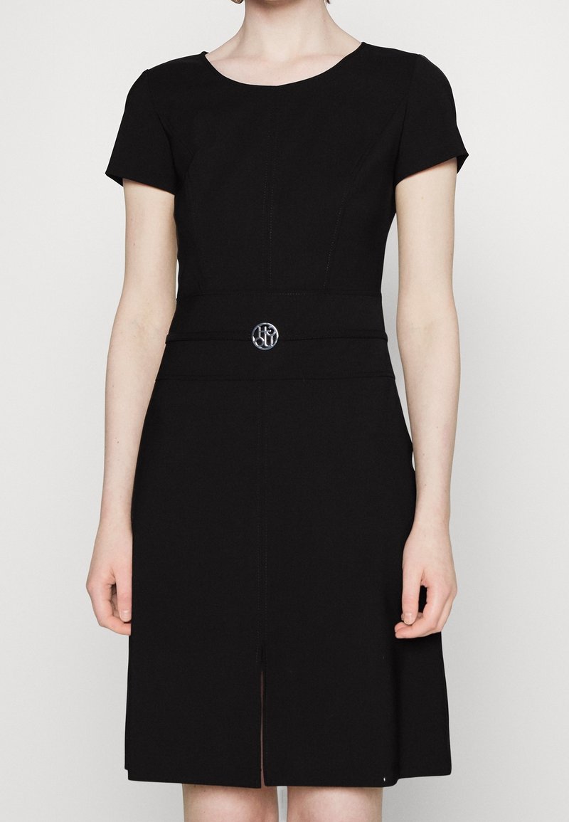 Woman wearing a knee-length black dress with short sleeves and a round neckline, featuring a decorative silver circular belt buckle.