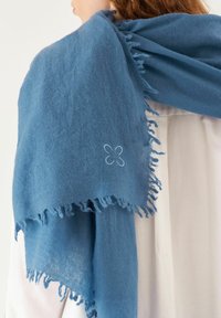 Écharpe bleue à franges en matériau doux, avec un détail de fleur brodé d'un côté. Drapée sur un vêtement clair.