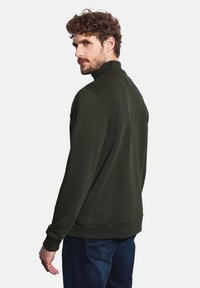 Tmavě zelená mikina s vysokým límcem, dlouhými rukávy a žebrovanými manžetami, spárovaná s modrými džíny, viděná zezadu.