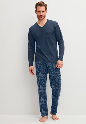 Man staat op blote voeten, draagt een marineblauwe top met lange mouwen en V-hals en blauwe pyjamabroek met patroon tegen een effen witte achtergrond.