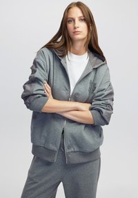 Sudadera gris con cremallera y capucha, acentos de botones y puños acanalados. Presenta paneles laterales en una tela gris contrastante. Llevada sobre una camiseta blanca.