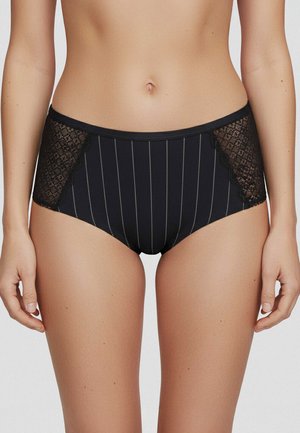 Kvinde iført sorte højtaljede trusser med lodrette striber og blondepaneler på hofterne, vist fra taljen til midt på låret.