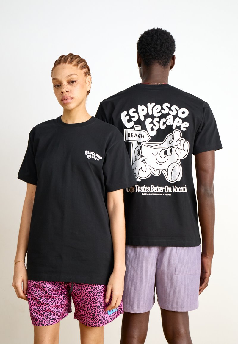 Svart bomull t-skjorte med et tegneserie-design på ryggen med tekst. Korte ermer og avslappet passform. Modellen har på seg rosa leopardmønstrede shorts.