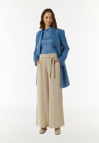 Blauwe op maat gemaakte jas over een lichtblauw topje, beige wijde broek met een strikband en huidkleurige hakken. Glad textiel.