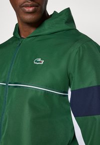 Zielona kurtka z kapturem wykonana z fakturowanej tkaniny; posiada biały pasek oraz ciemnoniebieski akcent na rękawie, z logo na piersi.