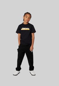 T-shirt noire avec logo DKNY blanc et jaune, assortie à un pantalon noir et des baskets blanches. Design simple et coupe décontractée.