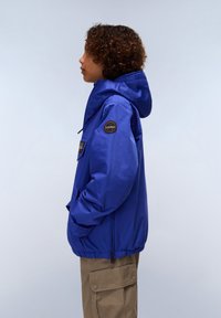 Modrá nepromokavá bunda s kapucí, obsahující přední kapsu, elastický lem a kulatou nášivku s logem na rukávu. V kombinaci s béžovými cargo kalhotami.