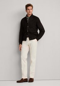 Homme portant une veste marron foncé boutonnée, une chemise verte, un pantalon couleur crème et des mocassins marron, debout devant un mur clair uni.