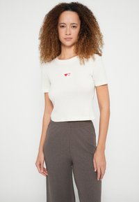 Crèmekleurig geribd T-shirt met korte mouwen en een cropped pasvorm, met twee rode hartborduursels op de borst. Gecombineerd met textuurrijke bruine broek met wijde pijpen.