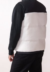 Padded vest s černým pleteným horním dílem a bílou plyšovou dolní částí. Disponuje kulatým výstřihem a manžetami. Je vidět texturovaný povrch.