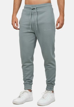 Lichtgrijze trainingsbroek met elastische tailleband en trekkoord, met ribgebreide boorden en subtiele branding op de dij. Draag met witte sneakers.