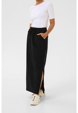 Femme portant un haut blanc à manches courtes, une jupe longue noire à cordon avec une fente latérale, et des baskets blanches épaisses, debout les mains dans les poches.