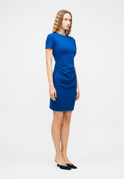 Γυναίκα με κυματιστά μαλλιά που φοράει εφαρμοστό φόρεμα μήκους μέχρι το γόνατο σε βασιλικό μπλε χρώμα και μαύρες γόβες με μυτερή μύτη, στέκεται μπροστά σε απλό άσπρο φόντο.