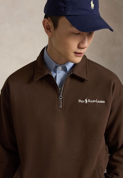 Νέος άνδρας φορούν καφέ πουλόβερ Polo Ralph Lauren με φερμουάρ μέχρι το στήθος, μπλε πουκάμισο με κουμπιά από κάτω και μπλε σκουφάκι με λογότυπο σε λευκό χρώμα.