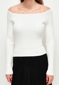 Weißes, schulterfreies, geripptes Strickoberteil mit langen Ärmeln und taillierter Silhouette, das mit einer strukturierten Oberfläche und einem figurbetonten Design versehen ist.