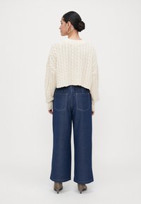 Nő, aki bunba tűzte a haját, krémszínű, cable-knit, rövid pulóverben, széles szárú sötétkék farmerben, hátsó zsebekkel, és taupe színű magassarkú cipőben áll.