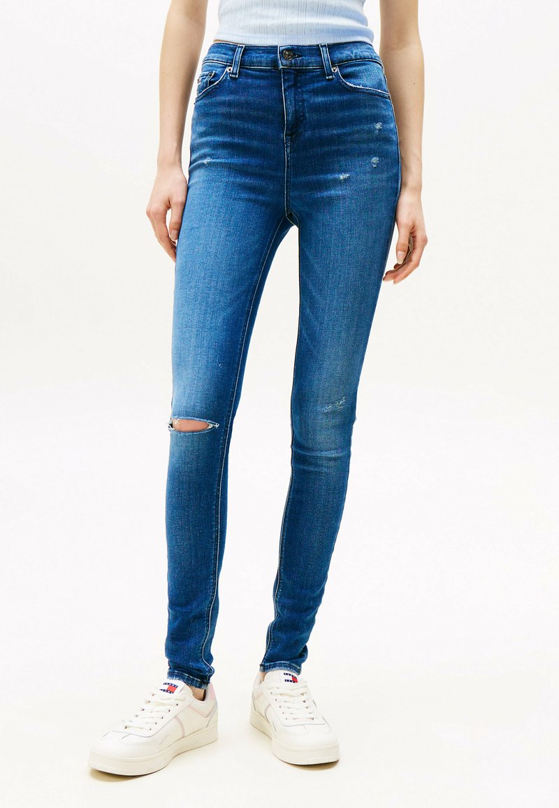Jeans de mezclilla azul ajustados con ligero desgastado, detalle de rodillas desgastadas y cierre de botón estándar. Combinados con zapatillas blancas.