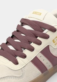 Close-up of a sneaker featuring cream leather, maroon laces, and two maroon stripes on the side, with a gold "GLOBE" logo.